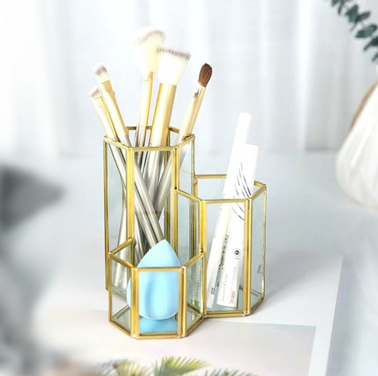 Gold and glass hexagonal makeup brush holder holding various brushes, a blue sponge, and white makeup pens.