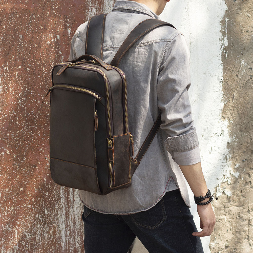 A man wears a brown, vintage-style leather backpack with multiple zippered compartments and sturdy shoulder straps.