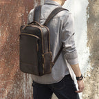 A man wears a brown, vintage-style leather backpack with multiple zippered compartments and sturdy shoulder straps.
