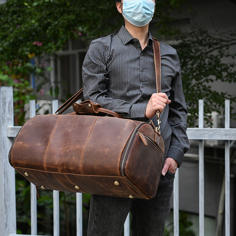 Man holding a vintage-style brown leather duffle bag with brass studs and a removable shoulder strap.
