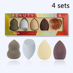Four makeup sponges in varied shapes/neutral tones and a rose-gold wire stand in a branded, colorful gift box.