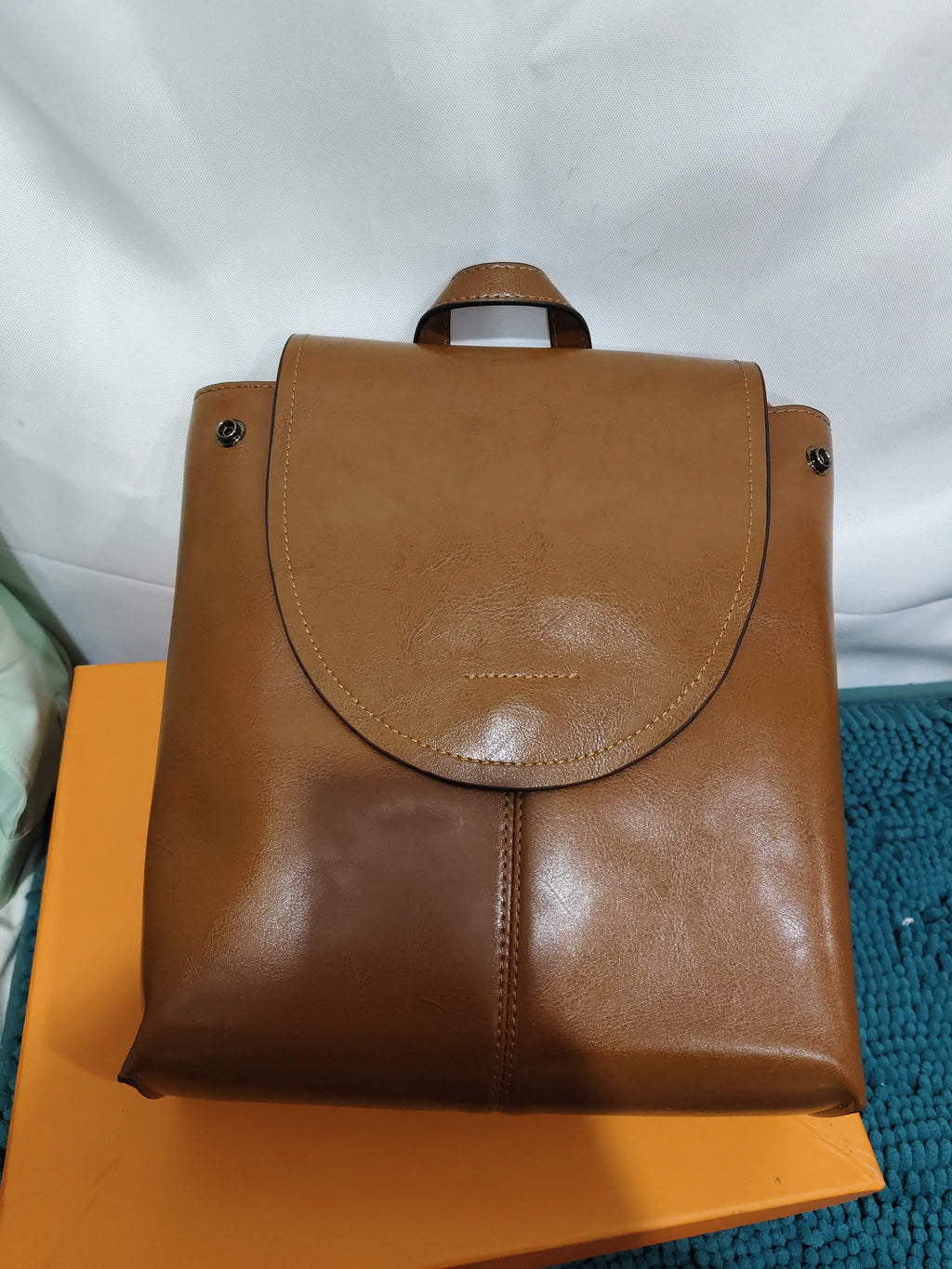 Tan leather women's retro messenger backpack with a flap top closure and top handle, resting on an orange box.
