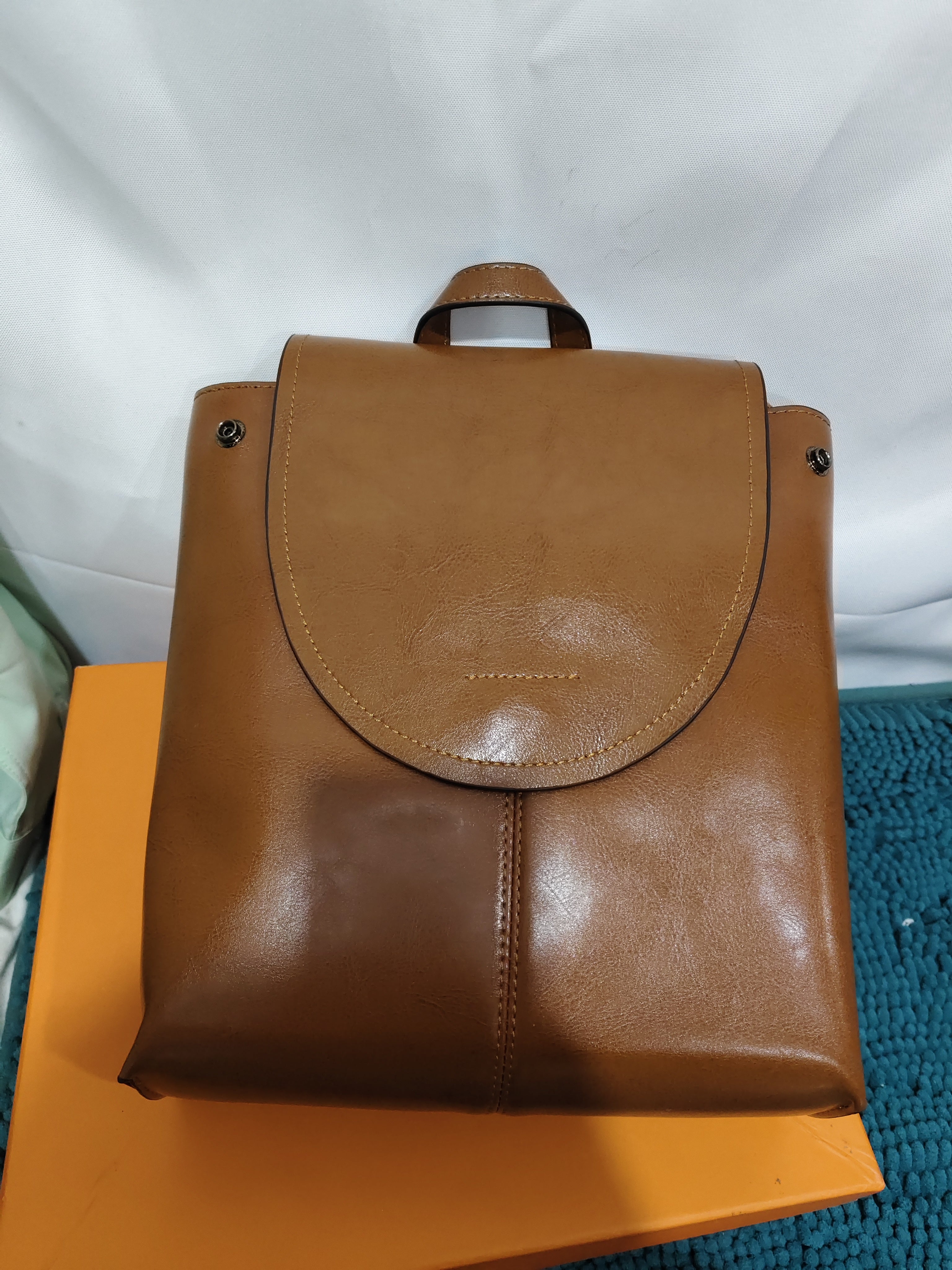 Tan leather women's retro messenger backpack with a flap top closure and top handle, resting on an orange box.