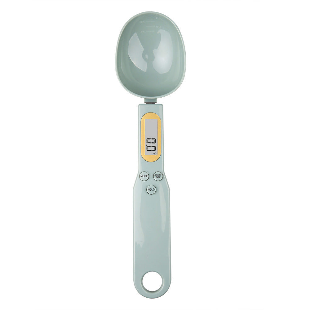 A sage green digital kitchen spoon scale with a yellow LCD display showing 0.0g and three control buttons.