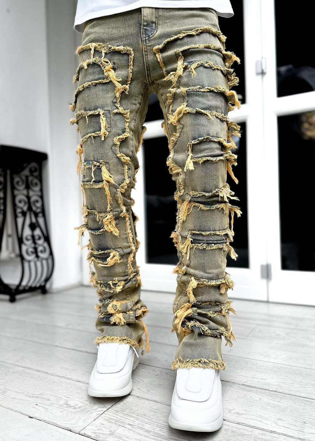 Men's stacked jeans in olive-toned denim featuring distressed, raw-edge fringe patches and a tight, distressed fit.