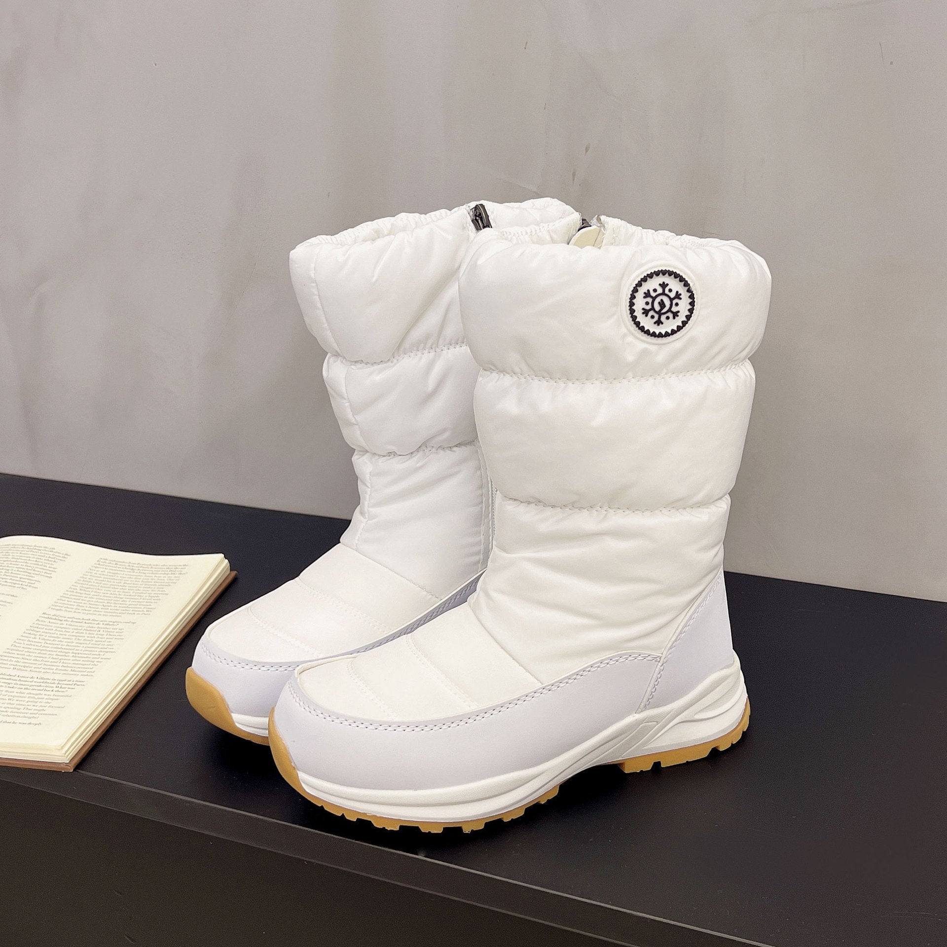White mid-calf snow boots with a quilted cotton upper, side zipper, circular snowflake logo, and tan lug soles.