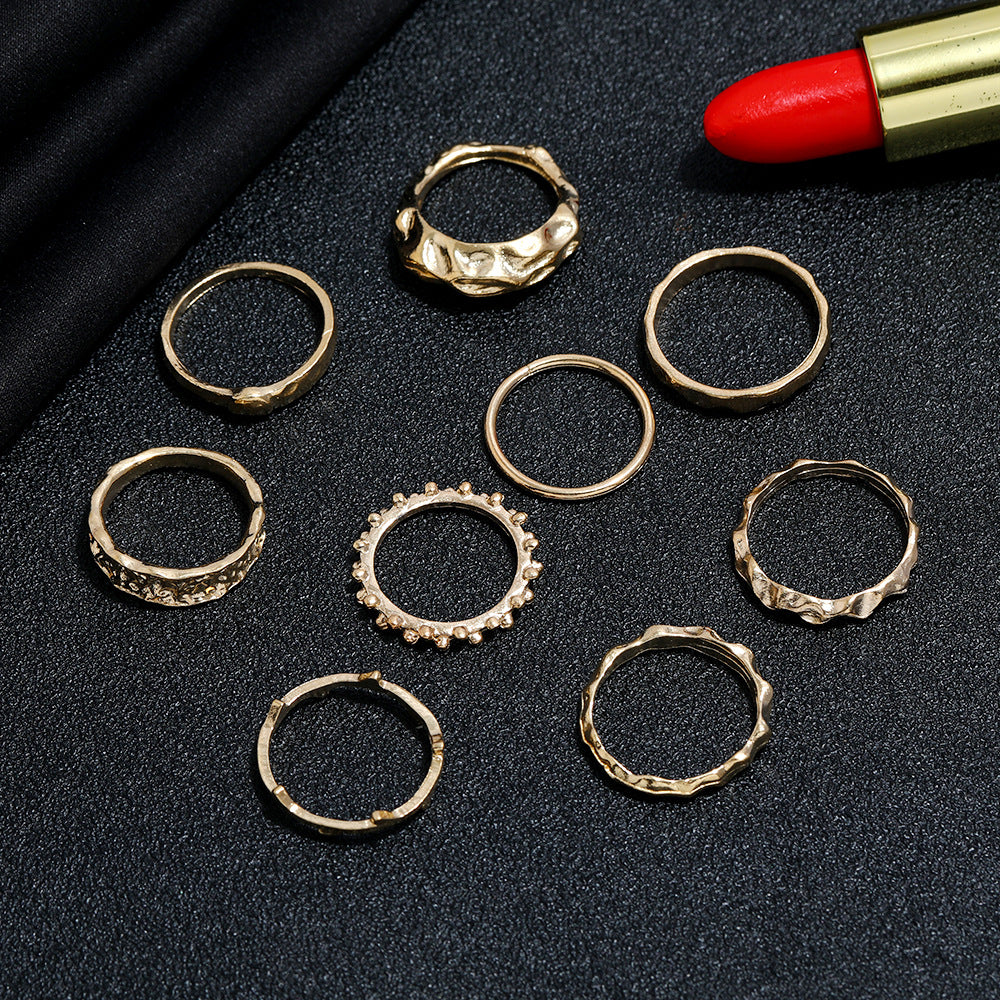 A set of eight varied gold-toned metal hip hop rings featuring textured and smooth minimalist designs on black fabric.