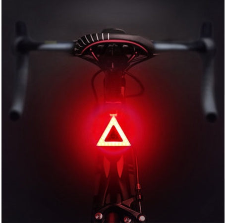 Bright red triangular LED bicycle taillight mounted under a black bike seat against a dark background.