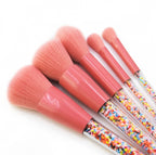 Five makeup brushes with soft pink bristles, pink metal ferrules, and clear handles filled with colorful candy sprinkles.