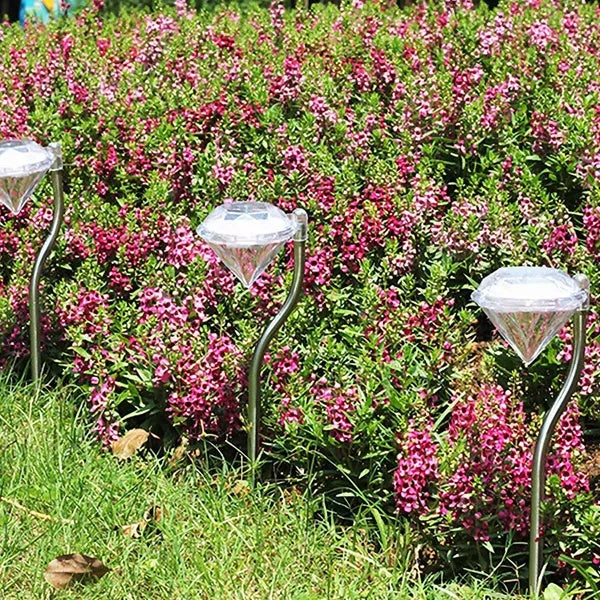 Four solar-powered diamond-shaped LED garden lights with silver stakes, placed in a garden with pink flowers and grass.