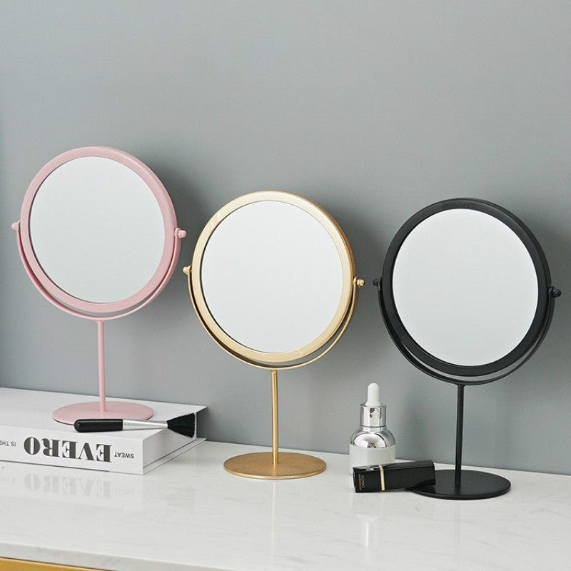 Three circular, rotating metal makeup mirrors in pink, gold, and black stand on a marble vanity surface.