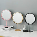 Three circular, rotating metal makeup mirrors in pink, gold, and black stand on a marble vanity surface.