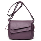 Soft purple leather crossbody bag with adjustable strap, overlapping flap design, and side zipper pull details.