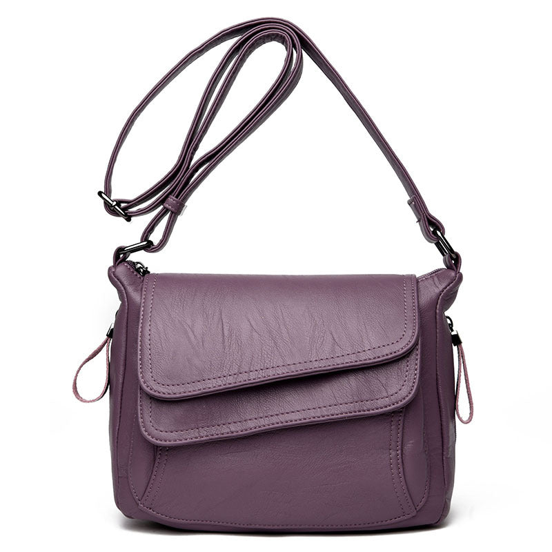 Soft purple leather crossbody bag with adjustable strap, overlapping flap design, and side zipper pull details.