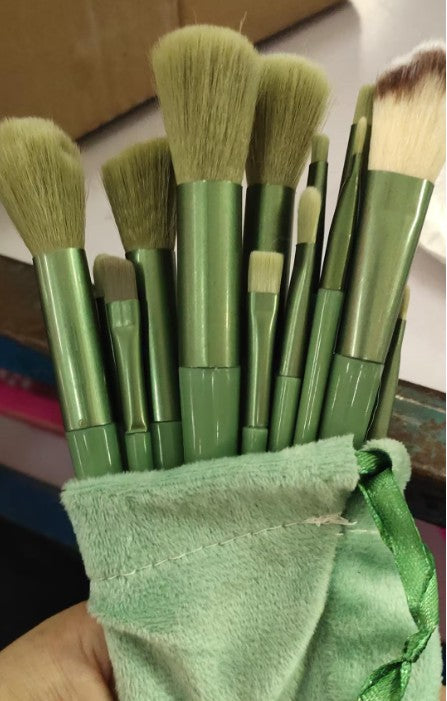 Set of 13 sage green makeup brushes with soft bristles, held in a matching velvet drawstring pouch.