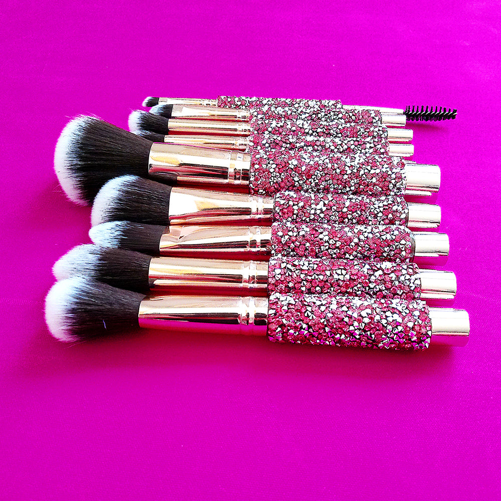 Set of 10 makeup brushes with gold ferrules and handles encrusted in sparkling pink and silver crystals on pink fabric.
