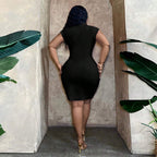 Back view of a woman wearing a fitted black V-neck mini dress with a cinched waist, standing between leafy green plants.