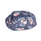 A dark grey, round, drawstring-style cosmetic bag with a pink and light blue floral pattern, closed with a front flap.