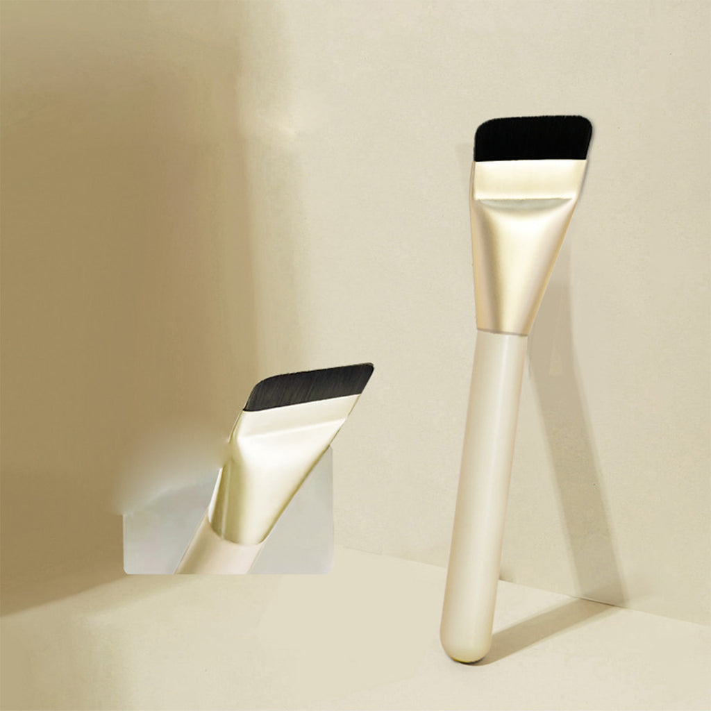 Ultra-thin makeup brush with black flat bristles, gold ferrule, and cream handle, shown in a clean, minimalist setting.