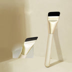 Ultra-thin makeup brush with black flat bristles, gold ferrule, and cream handle, shown in a clean, minimalist setting.