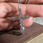 A silver-toned hip hop cross necklace featuring round, brilliant-cut clear diamonds, held against a hand.