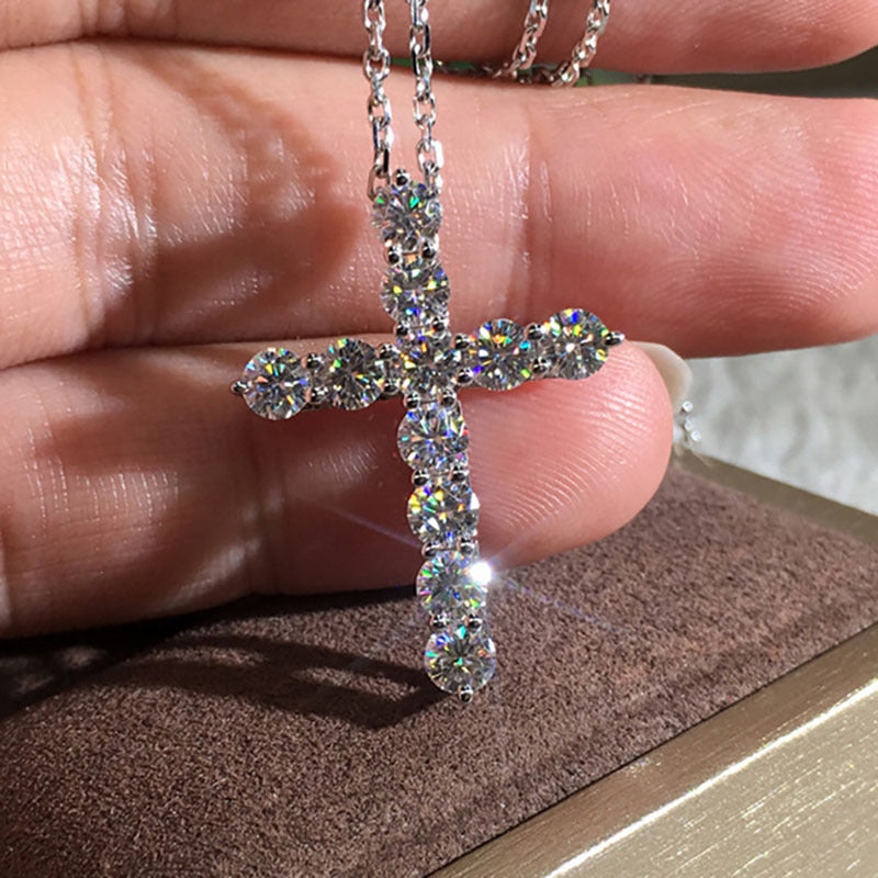 A silver-toned hip hop cross necklace featuring round, brilliant-cut clear diamonds, held against a hand.