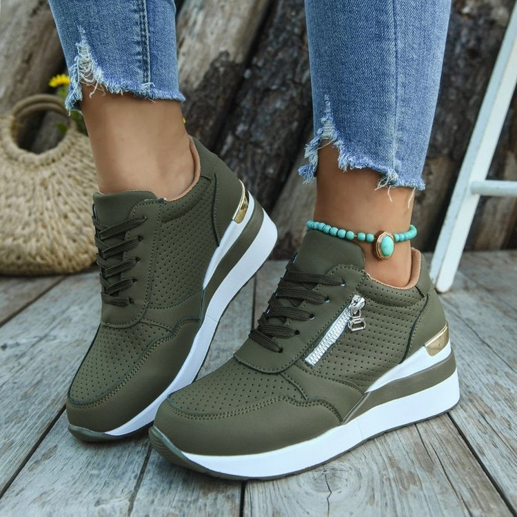 Olive green perforated sneakers with white platforms, laces, and side zippers worn with blue jeans and an anklet.