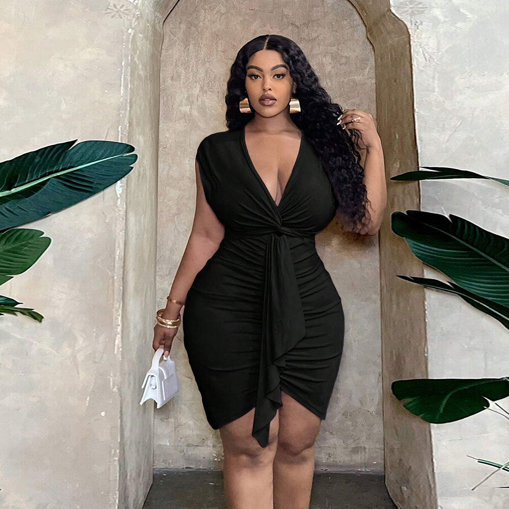 A woman wears a black, V-neck, ruched bodycon dress with a waist-tie and draped ruffle detail in a neutral setting.