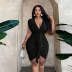 A woman wears a black, V-neck, ruched bodycon dress with a waist-tie and draped ruffle detail in a neutral setting.