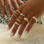 A hand wearing a set of gold-toned metal rings with varied textures, including smooth, hammered, and wave-like designs.