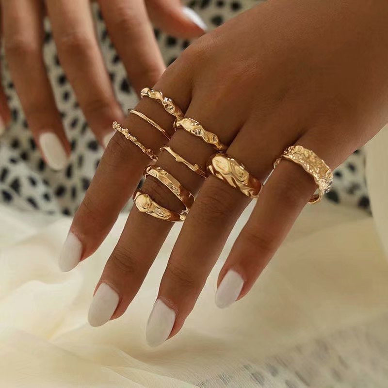 A hand wearing a set of varied gold-tone metal rings with textured, hammered, and smooth hip-hop style designs.