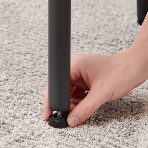 A hand adjusts the screw-in, adjustable black leveling foot on the metal leg of a bar stool on a grey carpet.