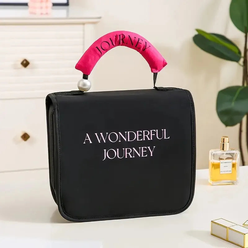 Black cosmetic bag with "A WONDERFUL JOURNEY" text, pink handle, and pearl accent, designed for travel.