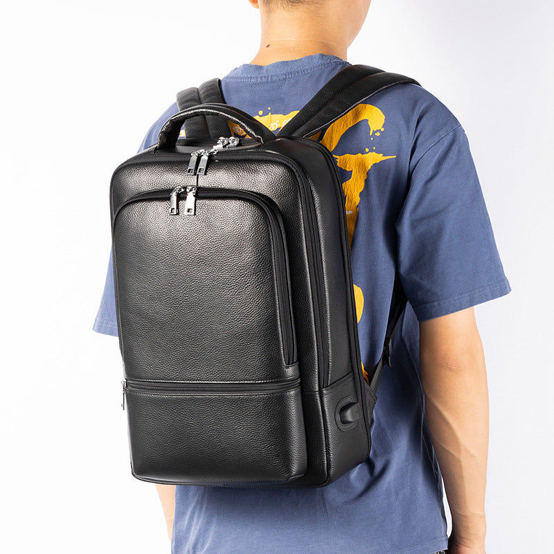 A man wears a black, textured leather business backpack with silver zippers, multiple compartments, and a USB port.