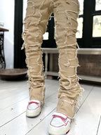 Tan stacked jeans with heavy distressed fringe detailing, featuring a slim fit and raw-edge patched construction.