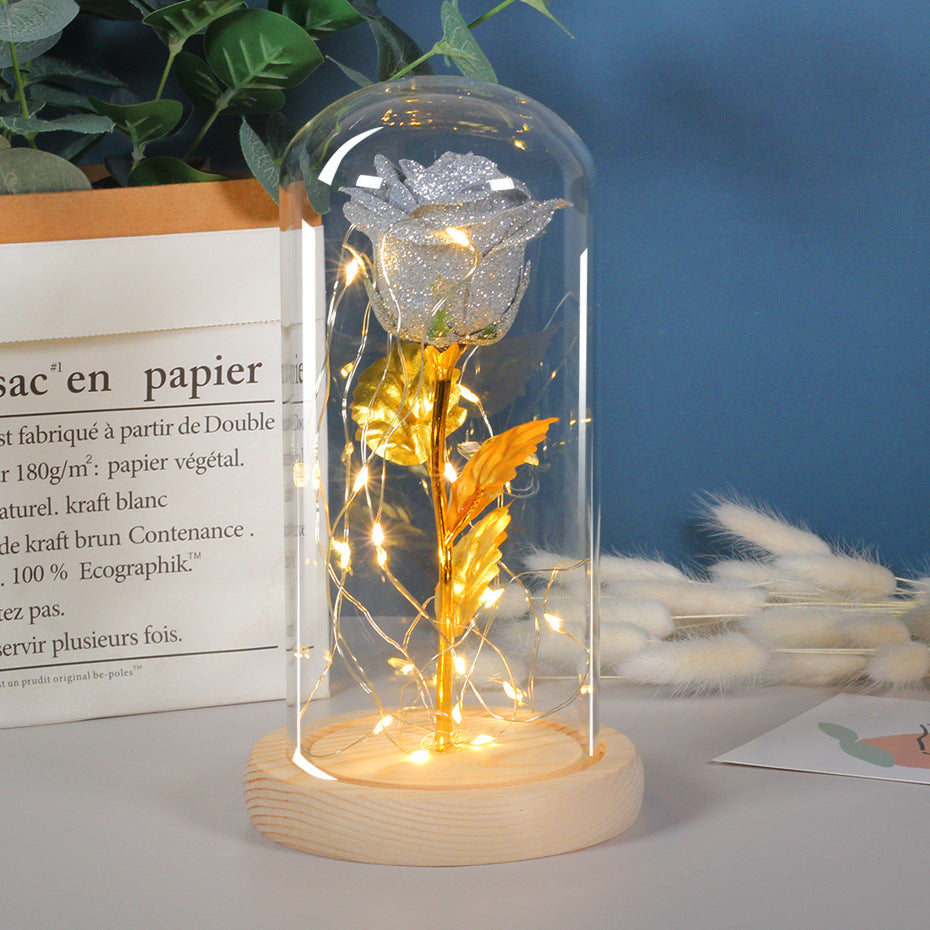 Silver glitter rose with gold stem inside a glass dome on a wooden base, illuminated by warm LED string lights.