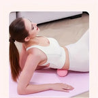 Woman in white activewear uses a small, pink, textured vibration massage ball on her lower back on a yoga mat.