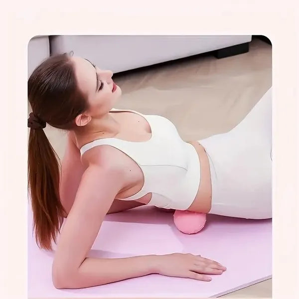 Woman in white activewear uses a small, pink, textured vibration massage ball on her lower back on a yoga mat.