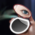 A hand holds a portable, round, dusty-rose LED compact makeup mirror reflecting an eye.