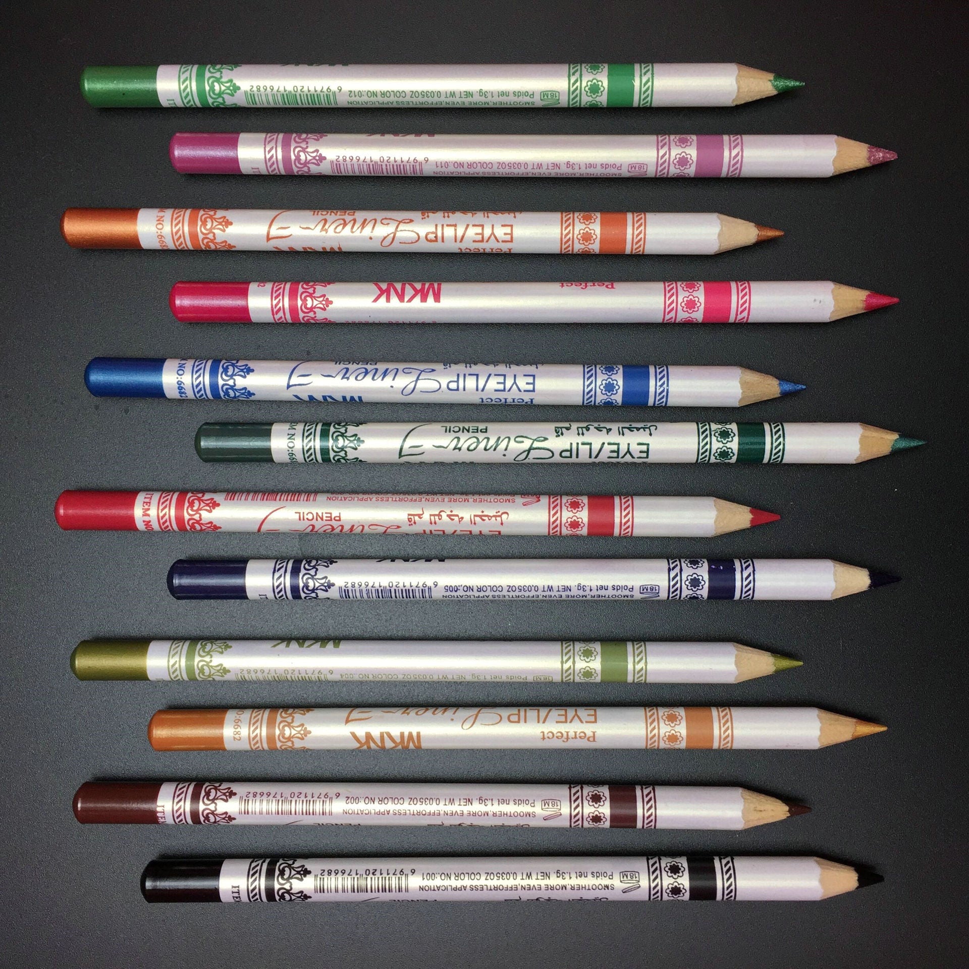Twelve glittery eye and lip liner pencils in various colors arranged horizontally against a dark background.