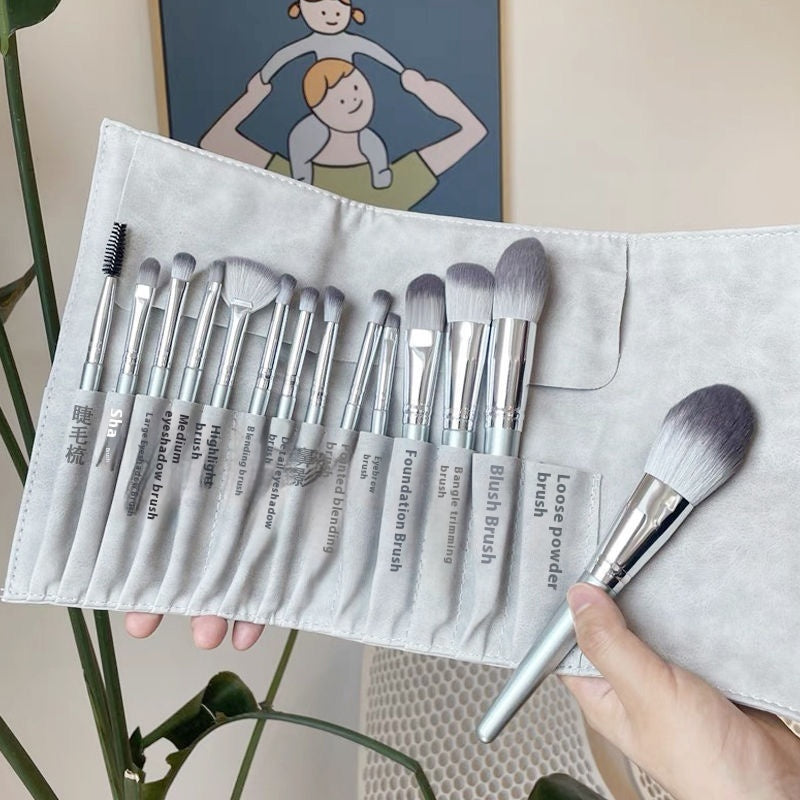 A gray suede roll-up case holding a 12-piece set of makeup brushes with silver ferrules and soft, two-toned bristles.