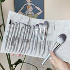 A gray suede roll-up case holding a 12-piece set of makeup brushes with silver ferrules and soft, two-toned bristles.