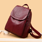 Maroon faux leather backpack with fold-over flap, embossed logo, and adjustable straps on a beige background.
