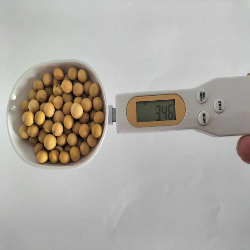 White digital kitchen spoon scale filled with soybeans, displaying a weight of 34.6 grams on its gold-toned LCD screen.