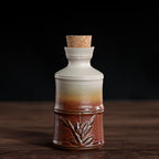 Ceramic wine decanter with a gradient cream-to-red glaze, bamboo-style texture, and a cork stopper on a wooden table.