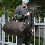 A man carries a brown, vintage-style leather duffle bag with a shoulder strap and side zipper pocket outdoors.