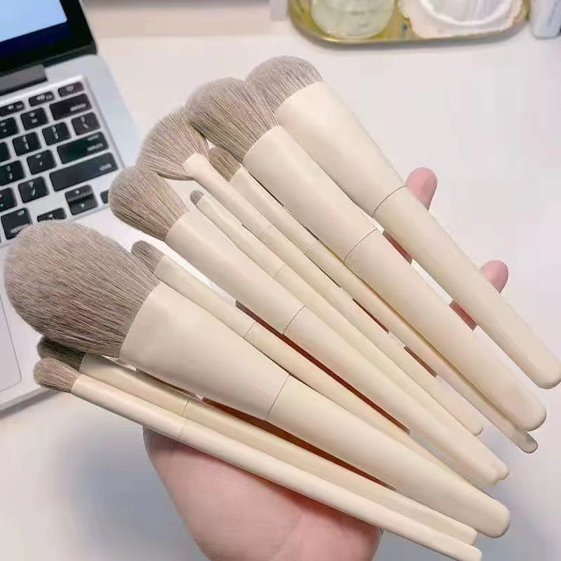 A hand holds a set of cream-colored makeup brushes with soft, light brown synthetic bristles against a laptop background.
