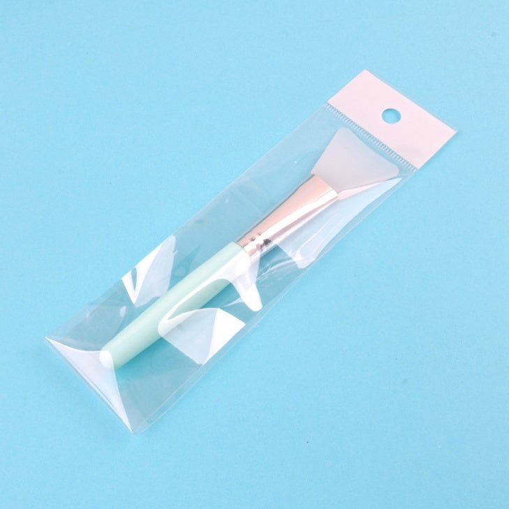 Silicone face mask brush in clear packaging, featuring a light green handle, rose gold ferrule, and white silicone head.