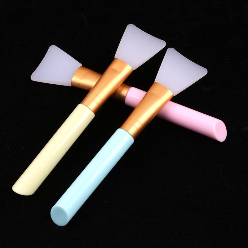 Three makeup brushes with white silicone heads, gold ferrules, and pastel yellow, blue, and pink handles on black.
