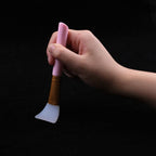 Hand holding a silicone facial mask brush with a pink handle, gold ferrule, and flexible white silicone head.